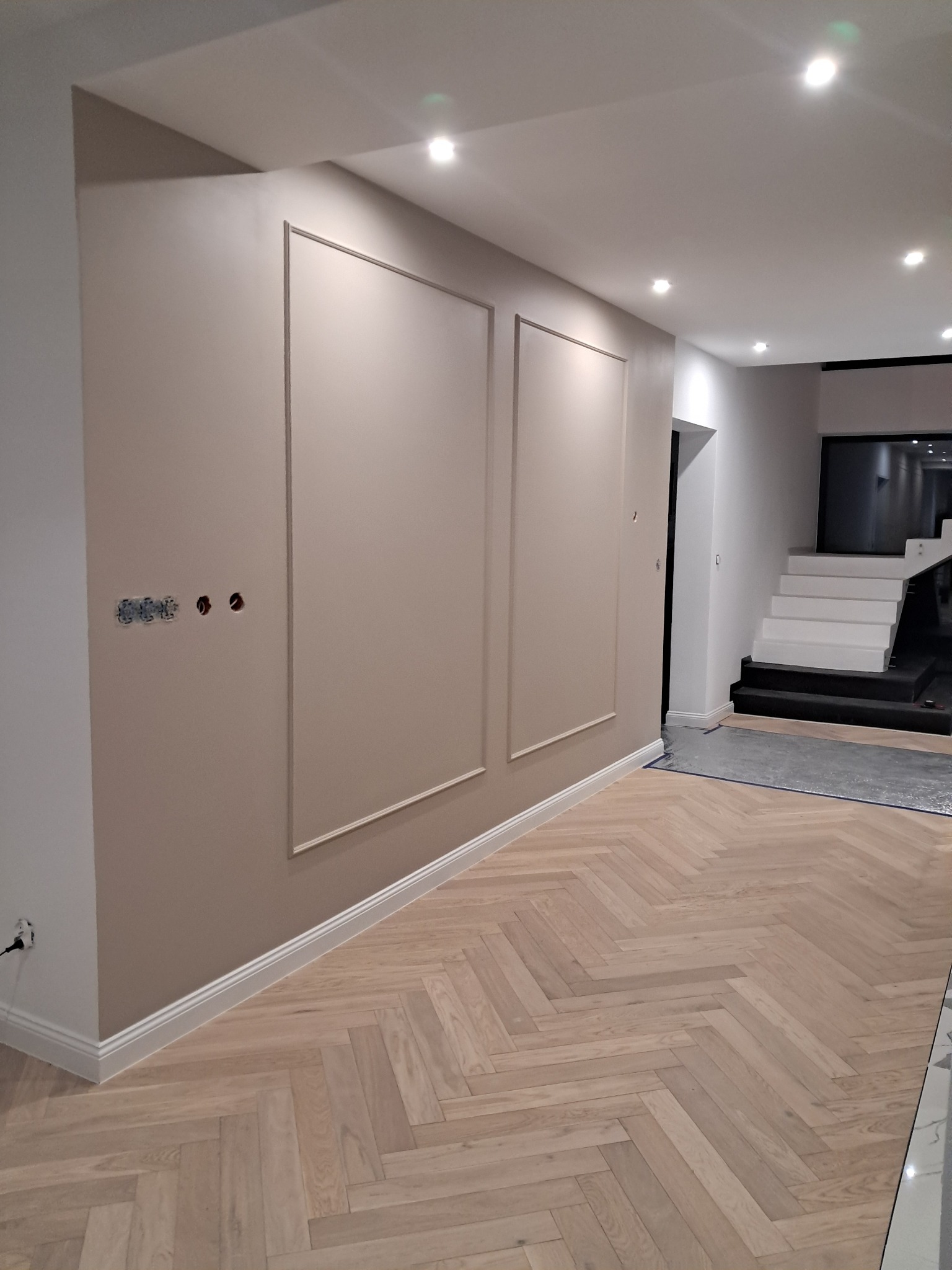 Beżowa ściana z ozdobnymi ramami i instalacją elektryczną, parkiet w jodełkę, białe listwy przypodłogowe i schody w tle. Nowoczesne wykończenie.