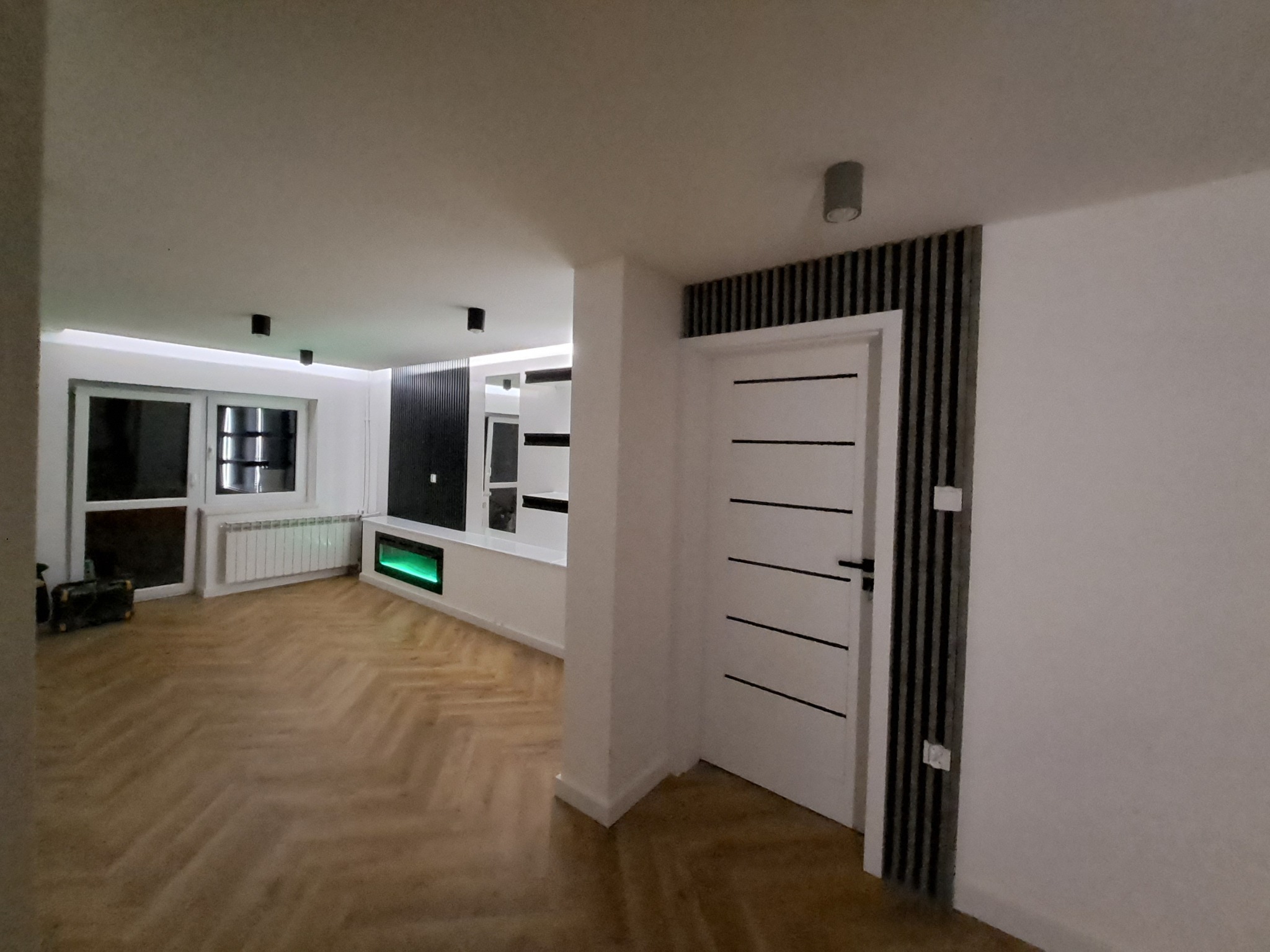 Salon po remoncie: parkiet w jodełkę, białe ściany, nowoczesne oświetlenie i drzwi z czarnymi akcentami. Widoczny fragment zabudowy z podświetleniem.