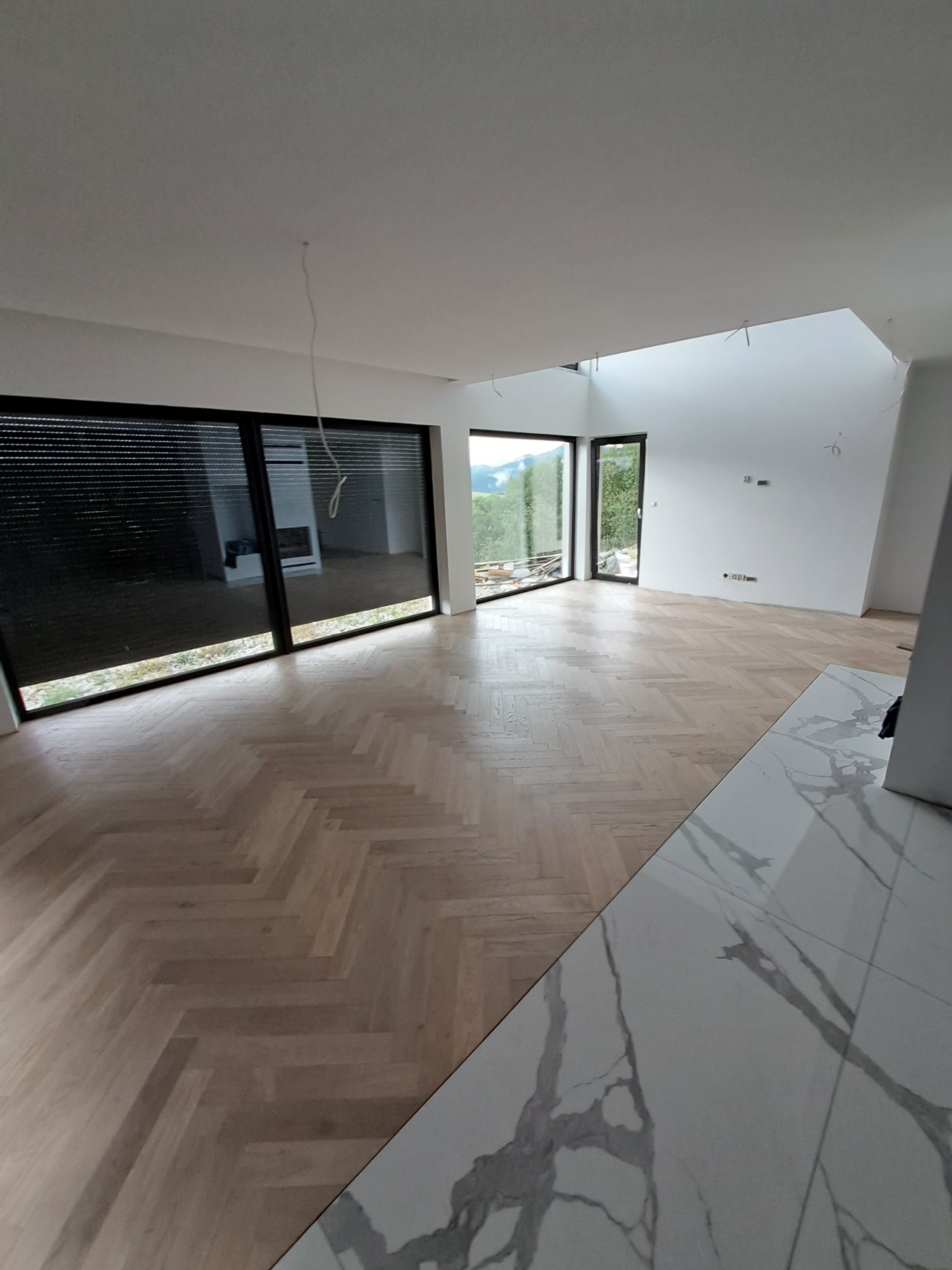 Jasne, parkietowe podłogi ułożone w jodełkę w przestronnym wnętrzu z dużymi oknami i marmurowymi płytkami. W tle widok na zieleń.