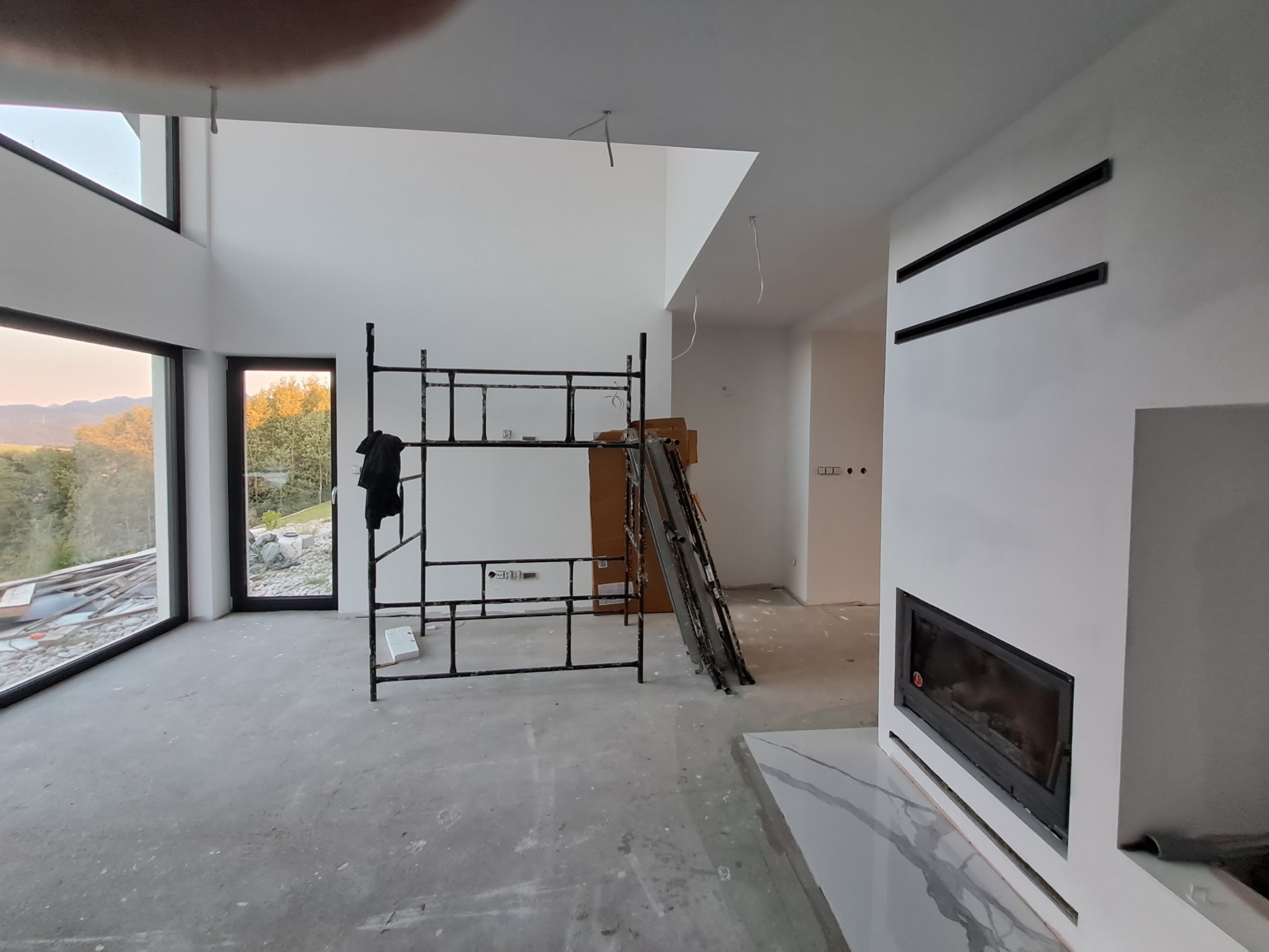 Wnętrze w trakcie wykończenia: białe ściany, duże okna, rusztowanie, kominek z czarną obudową. Widoczne elementy instalacji elektrycznej i surowa podłoga.