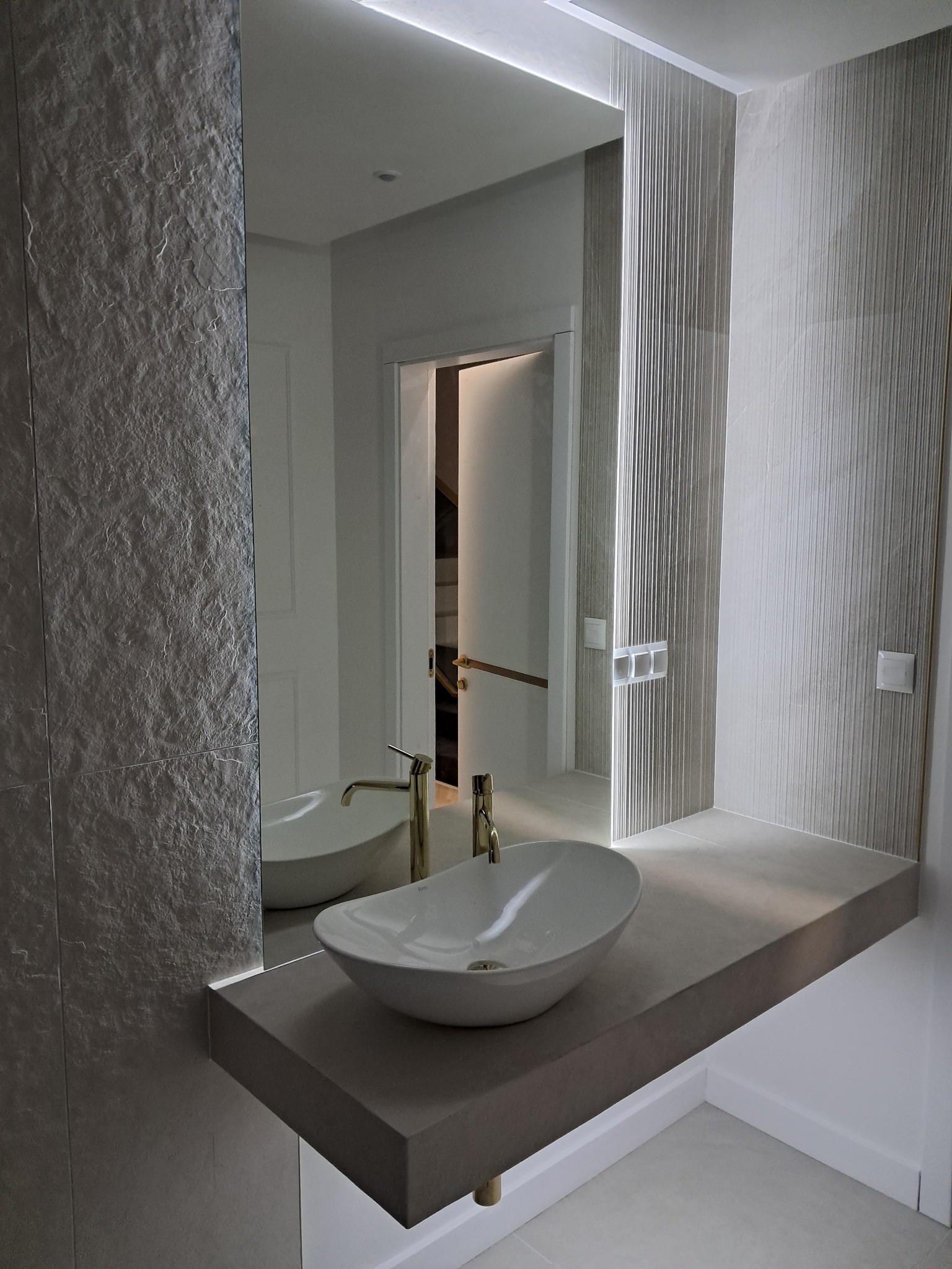 Nowoczesna łazienka z umywalką nablatową, złotą armaturą i podświetlanym lustrem. Po lewej ściana z szarej kamiennej okładziny, po prawej ściana z tapetą w pionowe prążki.