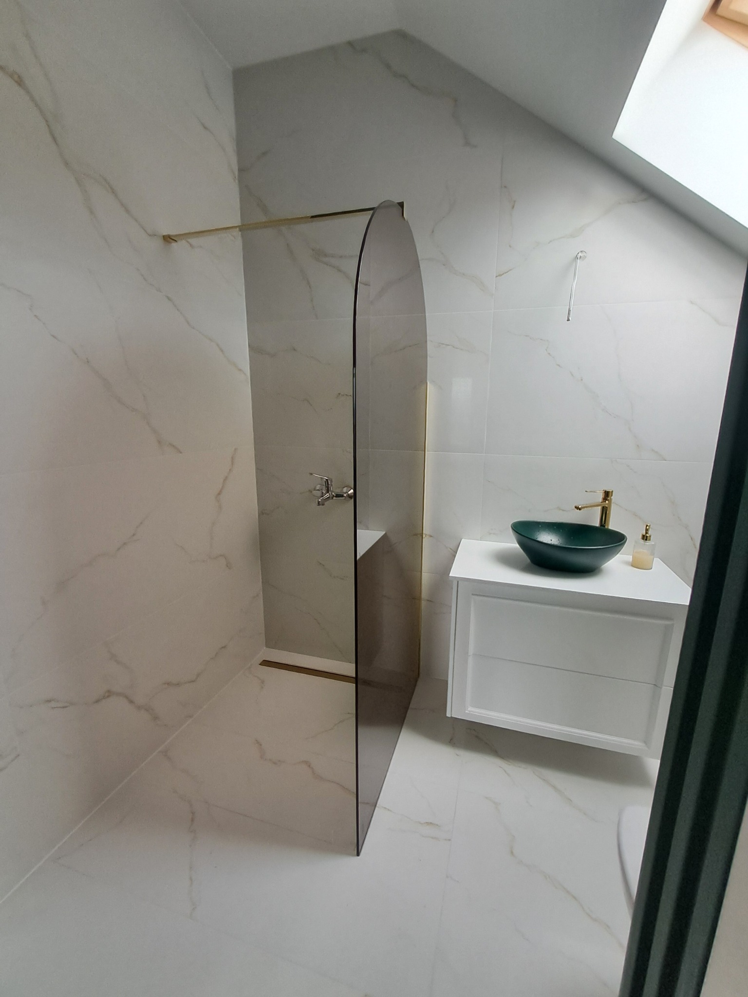 Nowoczesna łazienka na poddaszu z kabiną walk-in, umywalką w kolorze butelkowej zieleni i złotą armaturą na tle białych płytek imitujących marmur.