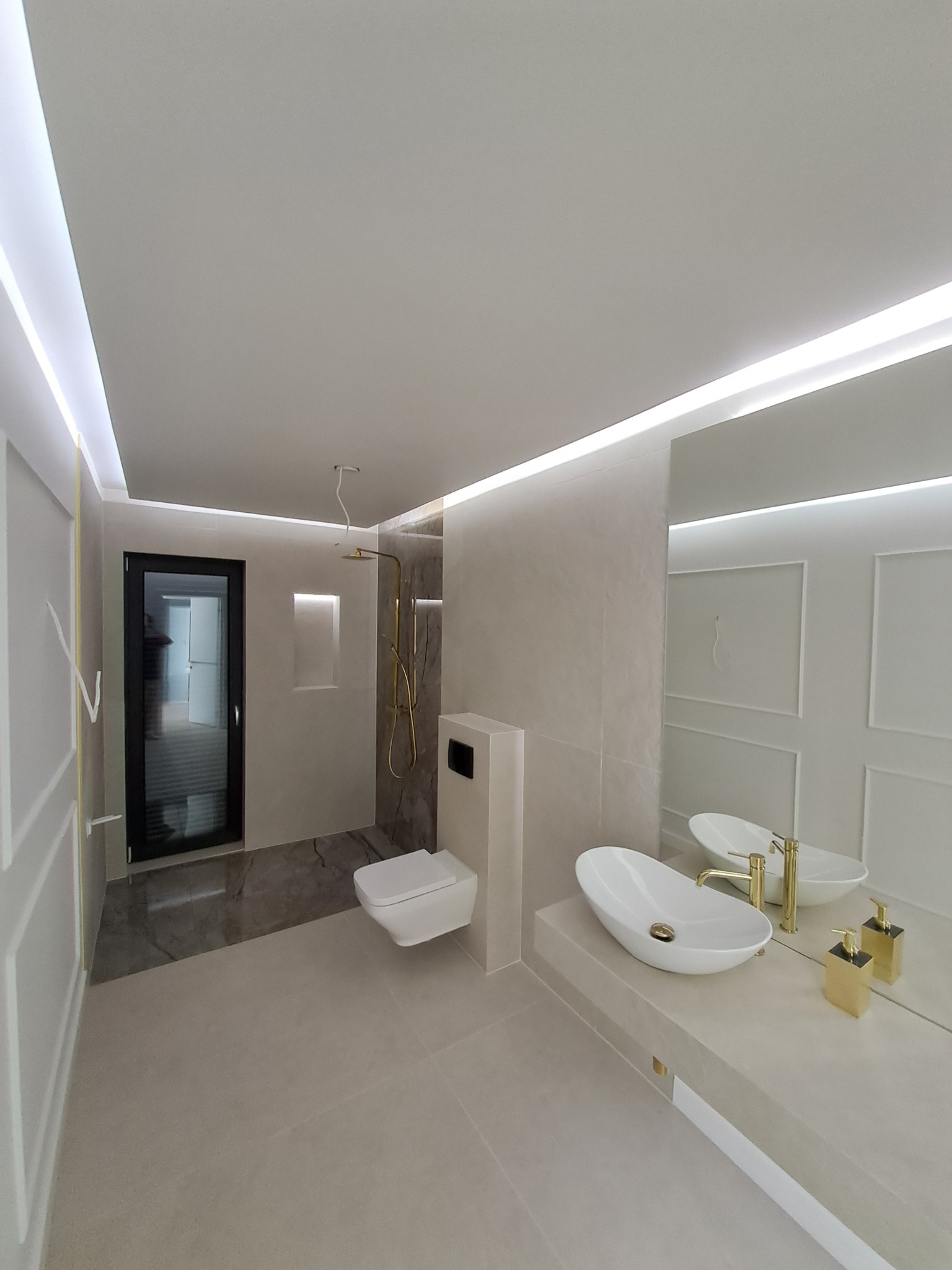 Nowoczesna łazienka z owalną umywalką, złotą armaturą i oświetleniem LED. Minimalistyczny design, jasne płytki, duże lustro i kabina prysznicowa typu walk-in.