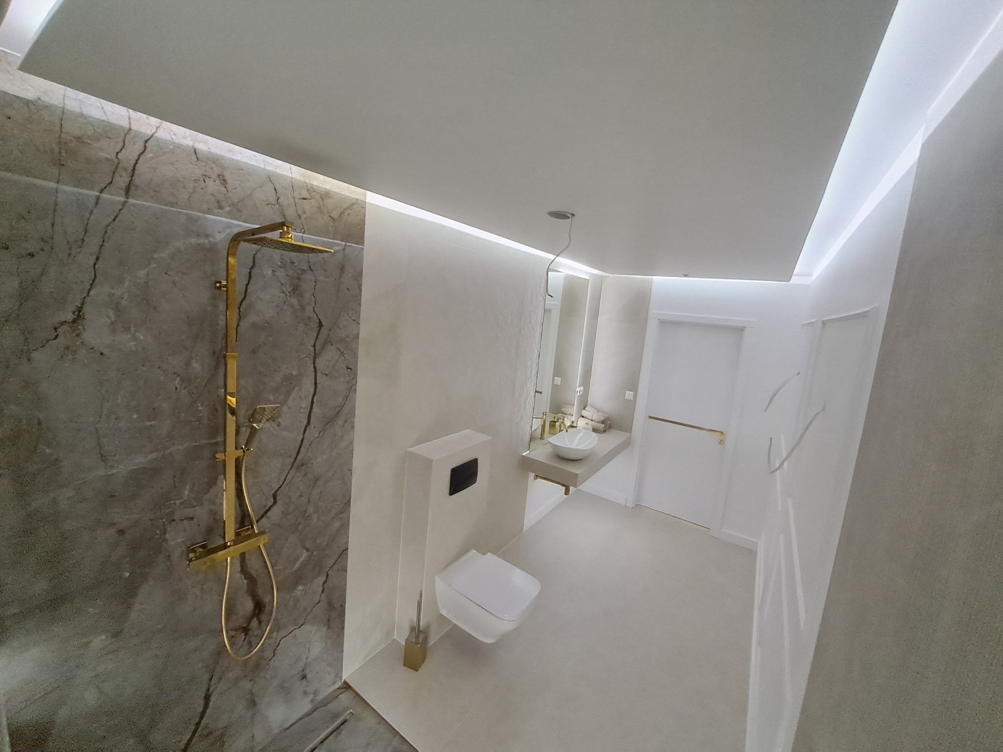 Wykończona łazienka w Nowym Targu: marmurowa ściana z złotą armaturą, minimalistyczna toaleta i umywalka na tle białych ścian z subtelnym oświetleniem.