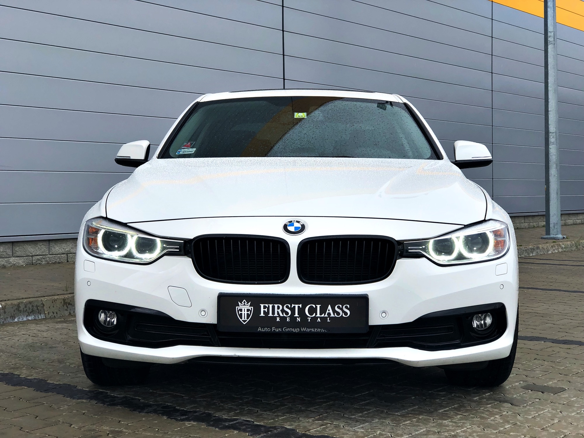 Przód białego BMW 320i z czarną atrapą chłodnicy i logo First Class Rental na tablicy rejestracyjnej, zaparkowanego na brukowanym placu przed szarym budynkiem z żółtym pasem.