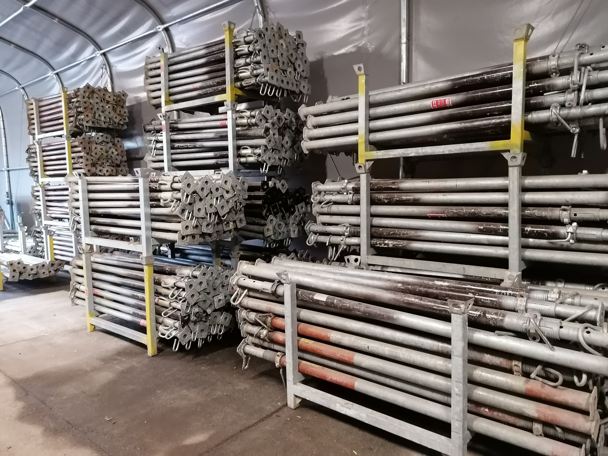 Magazynowane, metalowe stemple budowlane w metalowych regałach wewnątrz hali namiotowej.