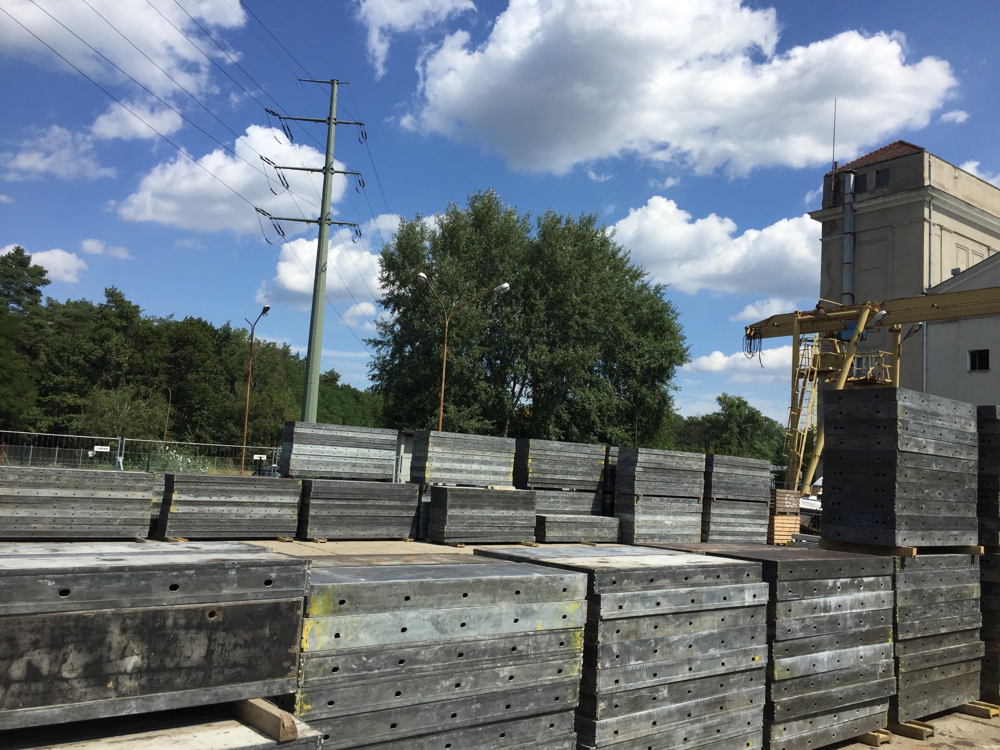 Stosy metalowych szalunków budowlanych na placu, widoczne ślady użytkowania, tło z błękitnym niebem i chmurami, słup wysokiego napięcia po lewej, fragment budynku z dźwigiem po prawej.