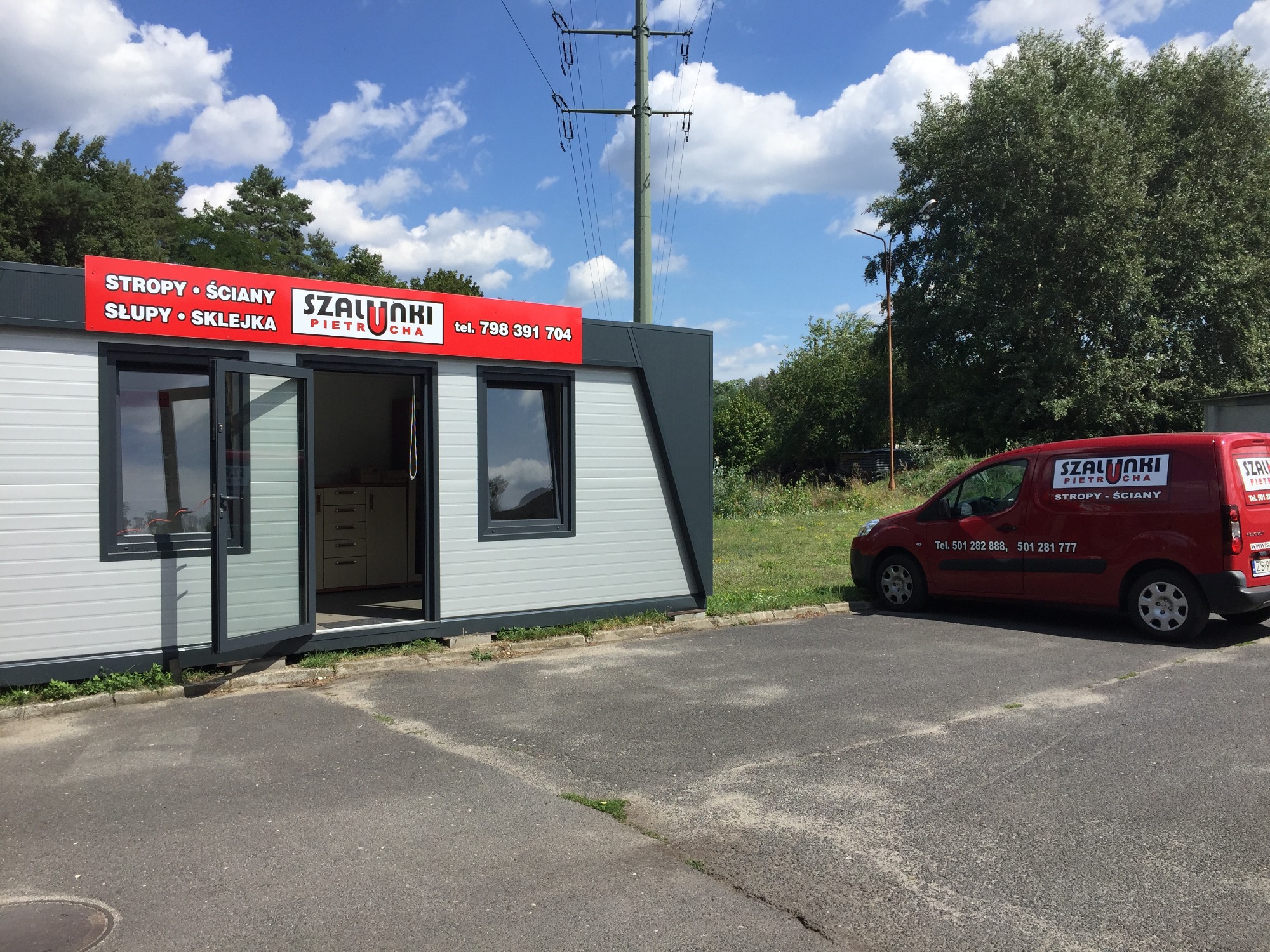 Kontener biurowy z otwartymi drzwiami, oznaczony szyldem z informacją o sprzedaży stropów, ścian, słupów i sklejki, obok zaparkowany czerwony samochód dostawczy z logo firmy i numerami telefonów...
