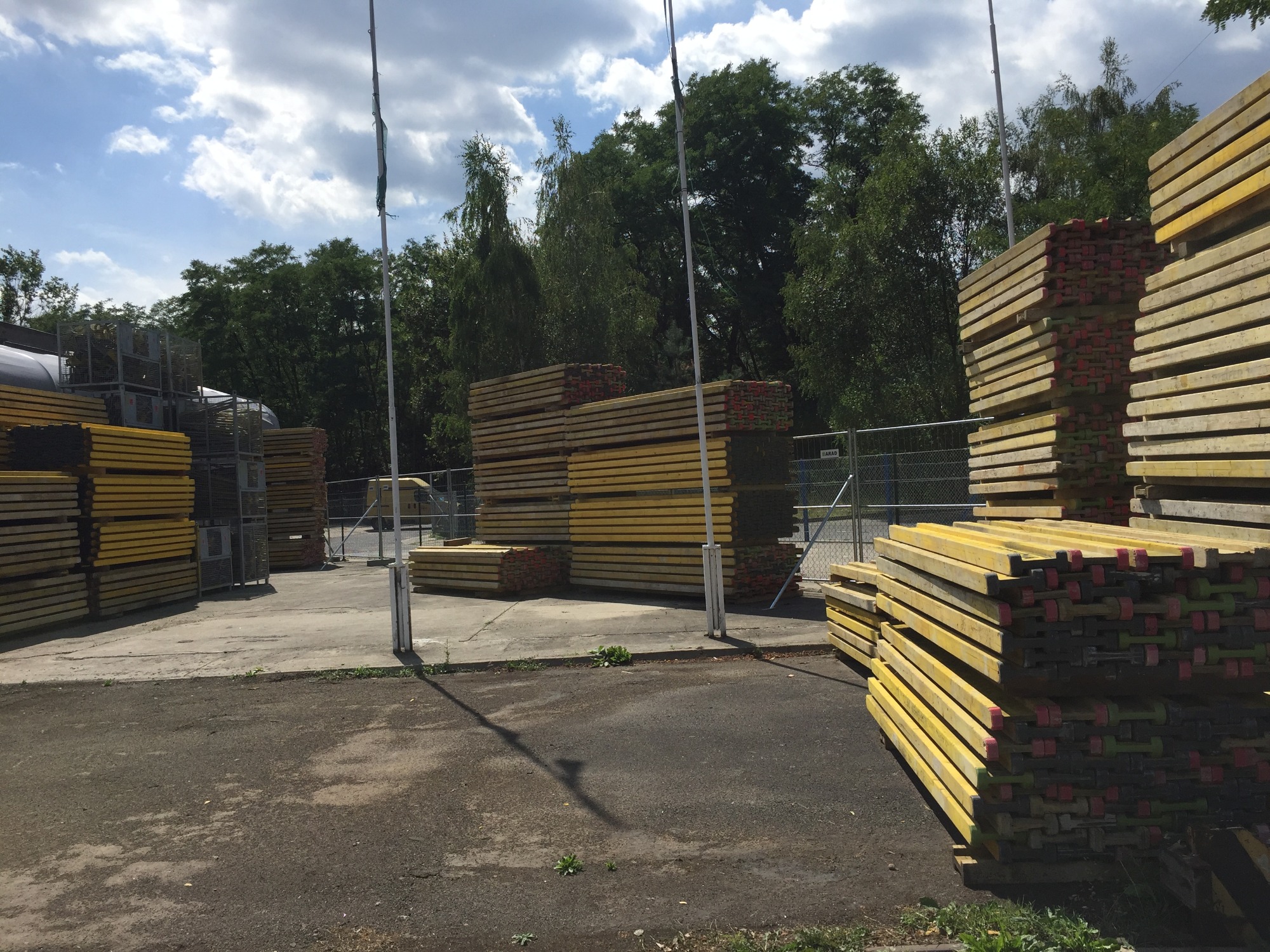 Stosy żółtych belek szalunkowych na placu budowy, widoczne zabezpieczenia i drzewa w tle, pochmurne niebo.