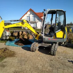 Żółto-szara minikoparka Wacker Neuson ET24 na kostce brukowej, z wykopaną ziemią obok i domami w tle, uchwycona w słoneczny dzień.