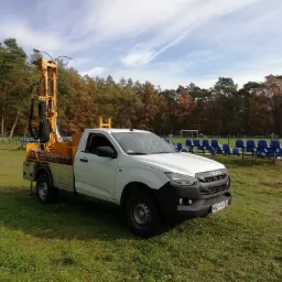 Biały pick-up Isuzu z żółtą wiertnicą zamontowaną na pace, stojący na trawie obok boiska sportowego z niebieskimi krzesełkami.