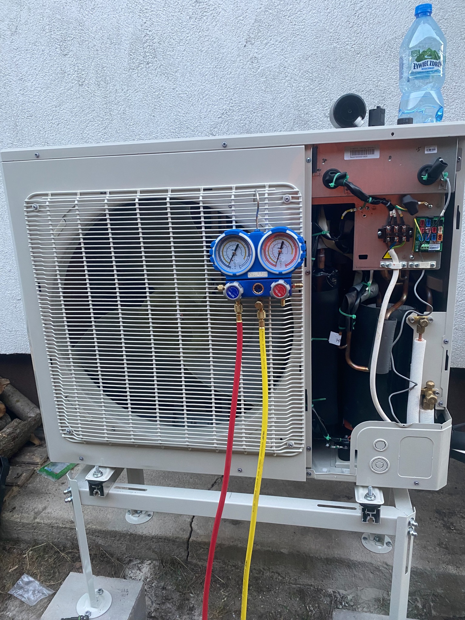 Klimatyzator zewnętrzny w trakcie serwisu z podłączonymi manometrami, widoczne przewody i elementy instalacji, butelka wody na obudowie.