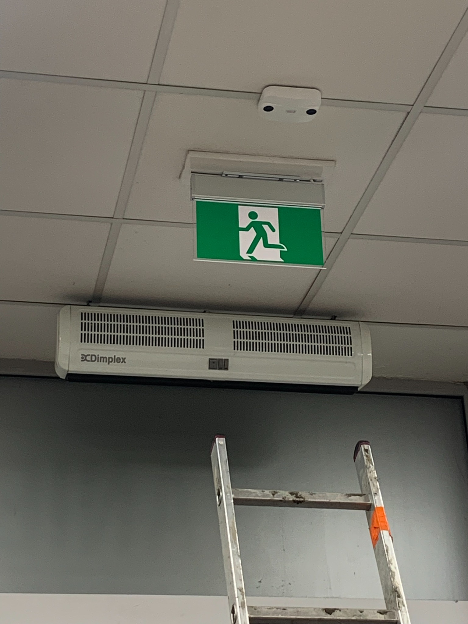 Biały panel grzewczy Dimplex zamontowany pod zielonym znakiem ewakuacyjnym na suficie kasetonowym, widoczny fragment aluminiowej drabiny.