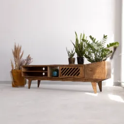 Drewniana szafka RTV w stylu retro z ukośnymi żaluzjami na drzwiczkach, z roślinami doniczkowymi na blacie i suszonymi trawami w torbie obok, na tle białej ściany.