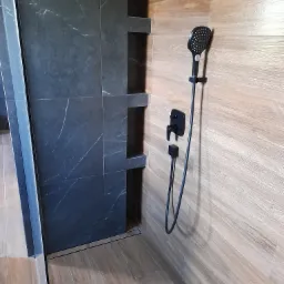 Kabina prysznicowa z czarnym panelem imitującym marmur, półkami wnękowymi i armaturą w kolorze czarnym, wykończona płytkami imitującymi drewno.