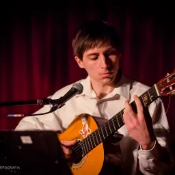 Młody mężczyzna w białej koszuli gra na gitarze klasycznej z naklejką tygrysa, mikrofon przed nim, czerwona zasłona w tle.