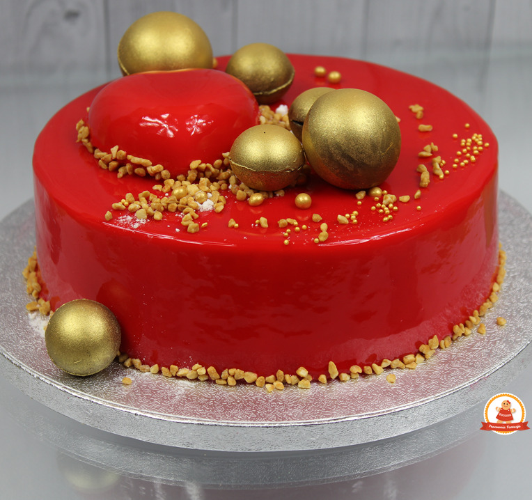 Tort z czerwoną, lustrzaną polewą, ozdobiony złotymi kulami i posypką z orzechów na srebrnej paterze.