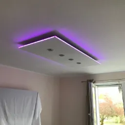 Podwieszany sufit z płyt kartonowo-gipsowych z wbudowanymi reflektorami i oświetleniem LED w kolorze fioletowym, widok z dołu, w tle okno z widokiem na drzewo.