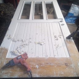 Paweł Koszyk - Zdjęcie przedstawia białe, drewniane drzwi z trzema szybkami, leżące poziomo na blacie, z częściowo usuniętą farbą. Widać opalarkę Black&Decker, używaną do usuwania starej farby, oraz ślady farby...