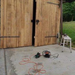 Paweł Koszyk - Drewniane, dwuskrzydłowe drzwi garażowe z metalowymi zawiasami, częściowo otwarte, przed nimi leżą dwa elektronarzędzia podłączone do sieci, obok stoi biały taboret.