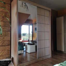 Paweł Koszyk - Szafa przesuwna z lustrem odbijająca wnętrze pokoju z wyjściem na balkon, widoczna część podłogi z paneli i dekoracyjna ściana z cegieł.