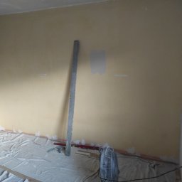 Paweł Koszyk - Pokój w trakcie przygotowania do malowania, z żółtą ścianą, częściowo zagruntowaną, zabezpieczoną podłogą i narzędziami malarskimi opartymi o ścianę.