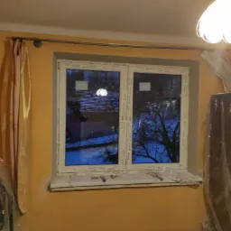 Pokój w trakcie remontu: pomarańczowe ściany, okno PCV z widokiem na drzewa, zasłonięte meble folią ochronną, widoczny karnisz i lampa sufitowa.