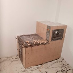 Paweł Koszyk - Demontaż kafli pieca kaflowego, widoczne fragmenty odspojonych płytek, zabezpieczona podłoga z narzędziami do demontażu w tle, ściany przygotowane do dalszych prac.