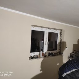 Paweł Koszyk - Montaż okna PCV w ścianie, widoczne narzędzia i piana montażowa, etap prac wykończeniowych w jasnym pomieszczeniu.
