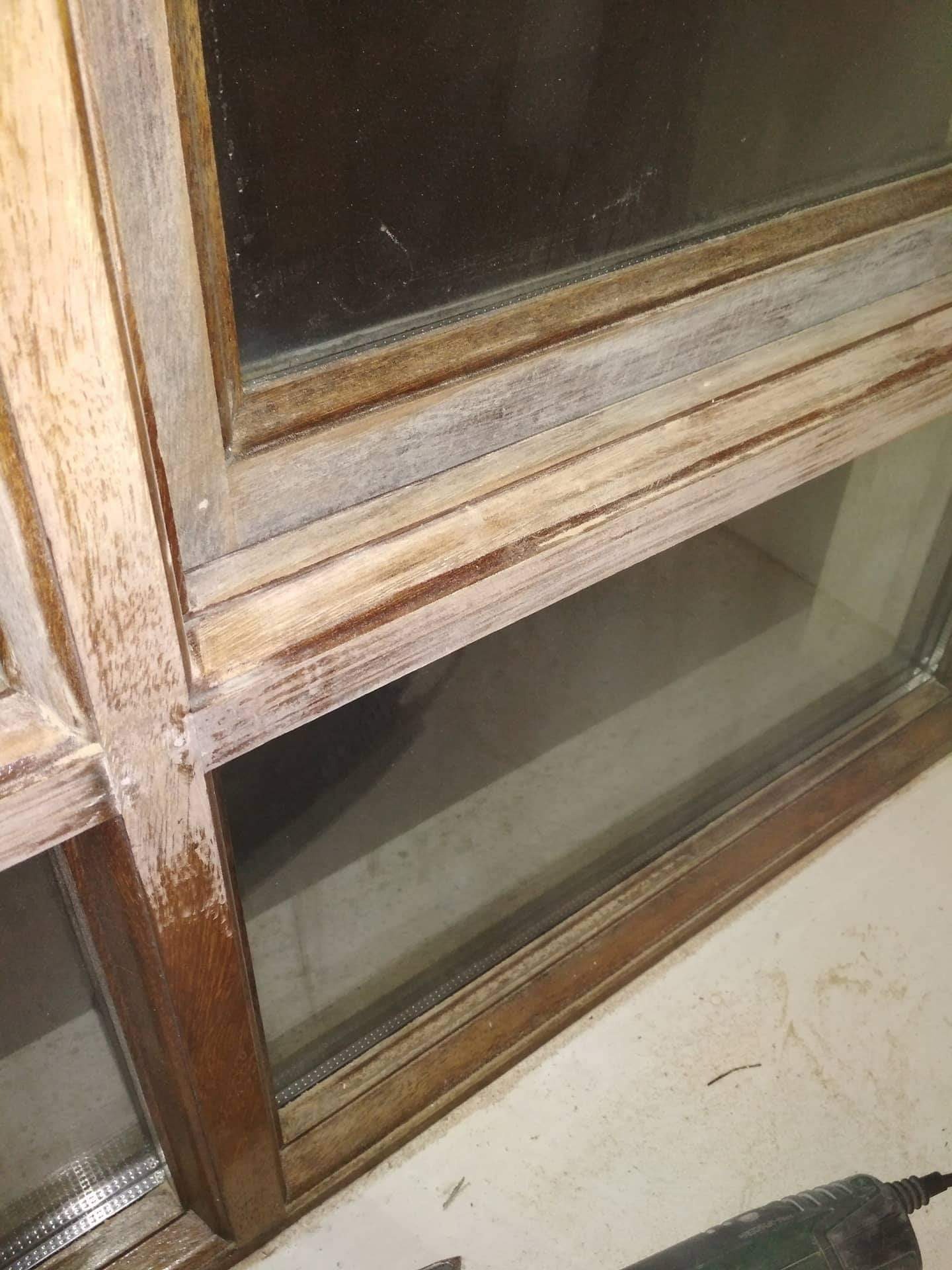 Fragment okna z drewnianą ramą, częściowo pokrytą białą farbą lub szpachlą, widoczne ślady po obróbce, w tle szyba i ściana, w dolnym rogu fragment szlifierki.