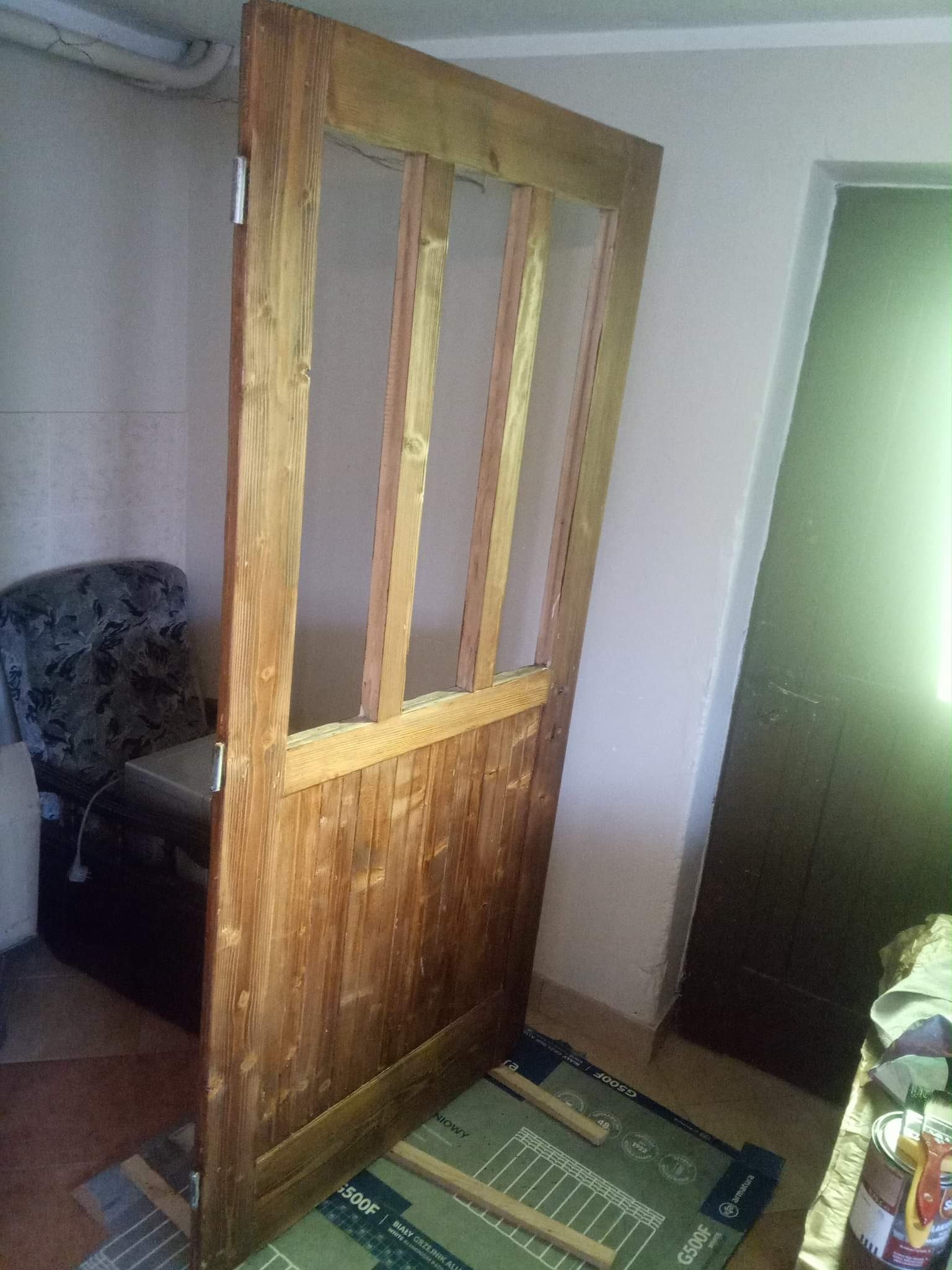 Drewniane drzwi wewnętrzne z szybami w trakcie renowacji, widoczne ślady farby i bejcy, na podłodze karton ochronny i akcesoria malarskie