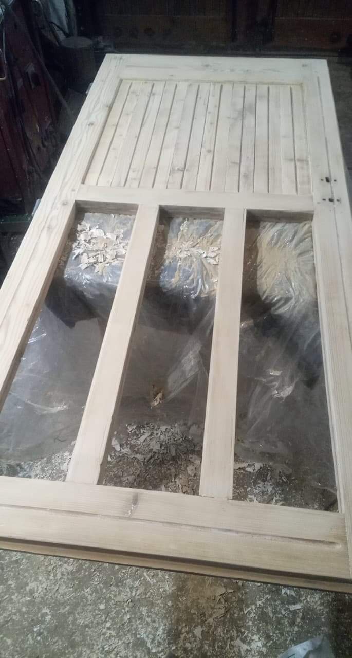 Surowa drewniana rama drzwi z trzema panelami w trakcie obróbki, leżąca na podłodze z widocznymi wiórami i folią ochronną.