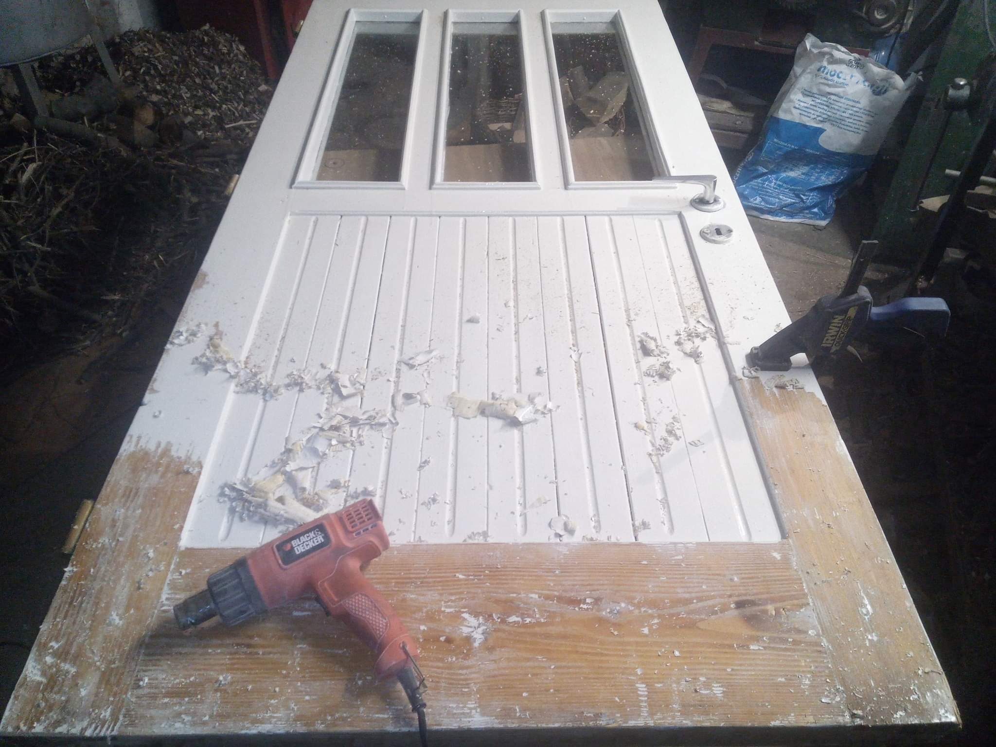 Zdjęcie przedstawia białe, drewniane drzwi z trzema szybkami, leżące poziomo na blacie, z częściowo usuniętą farbą. Widać opalarkę Black&Decker, używaną do usuwania starej farby, oraz ślady farby...