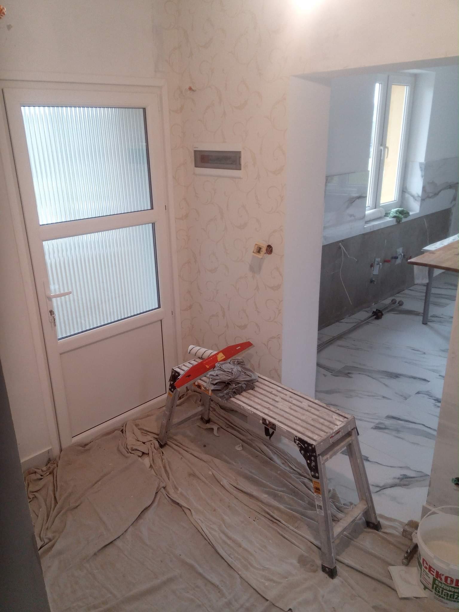 Pokój w trakcie remontu: widoczna drabina malarska z wałkiem i szpachelką, zakryta podłoga, ściana z tapetą w kwiatowy wzór, w tle okno i marmurowe płytki.