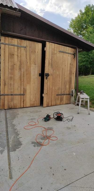 Drewniane, dwuskrzydłowe drzwi garażowe z metalowymi zawiasami, częściowo otwarte, przed nimi leżą dwa elektronarzędzia podłączone do sieci, obok stoi biały taboret.