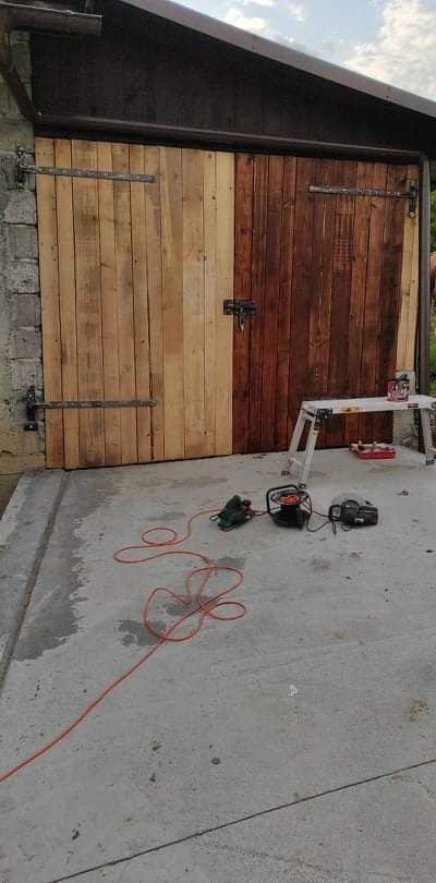 Drewniane wrota garażowe w trakcie odnawiania: jedna część surowa, druga po bejcowaniu, na podłodze szlifierka i przedłużacz, w tle drabina malarska.
