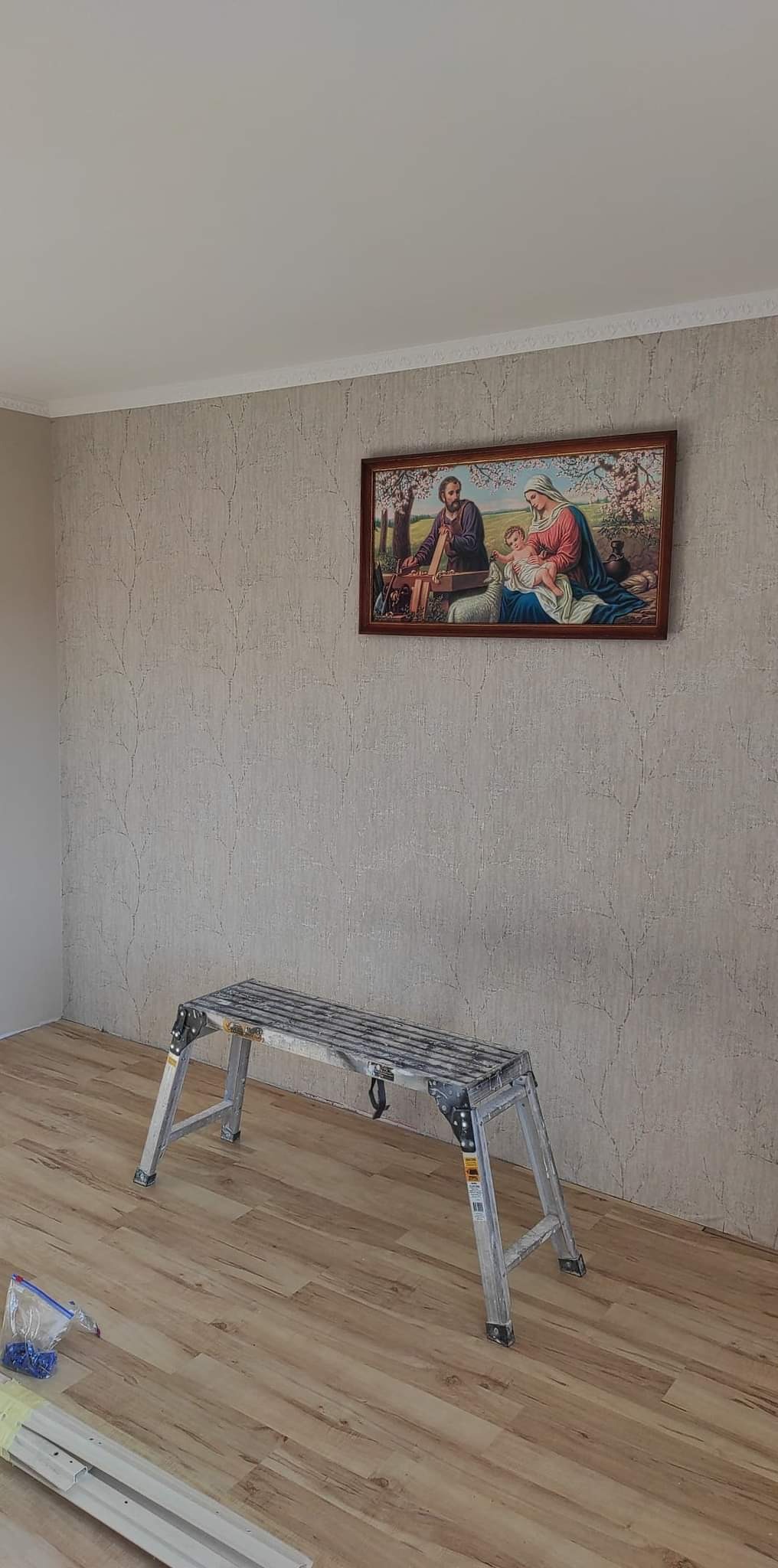 Pokój po remoncie z nową tapetą w odcieniach beżu, na ścianie wisi obrazek w drewnianej ramie, a na podłodze leżą panele oraz stoi aluminiowy podest.