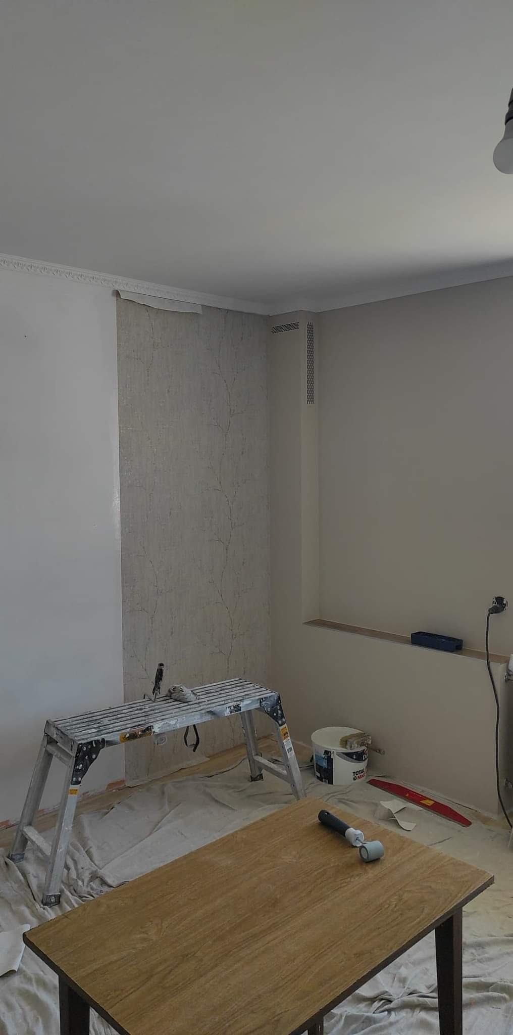 Fragment pokoju w trakcie tapetowania: częściowo położona tapeta w delikatny wzór drzewny, aluminiowa drabina malarska, wiadro z klejem i akcesoria na zabezpieczonej podłodze.