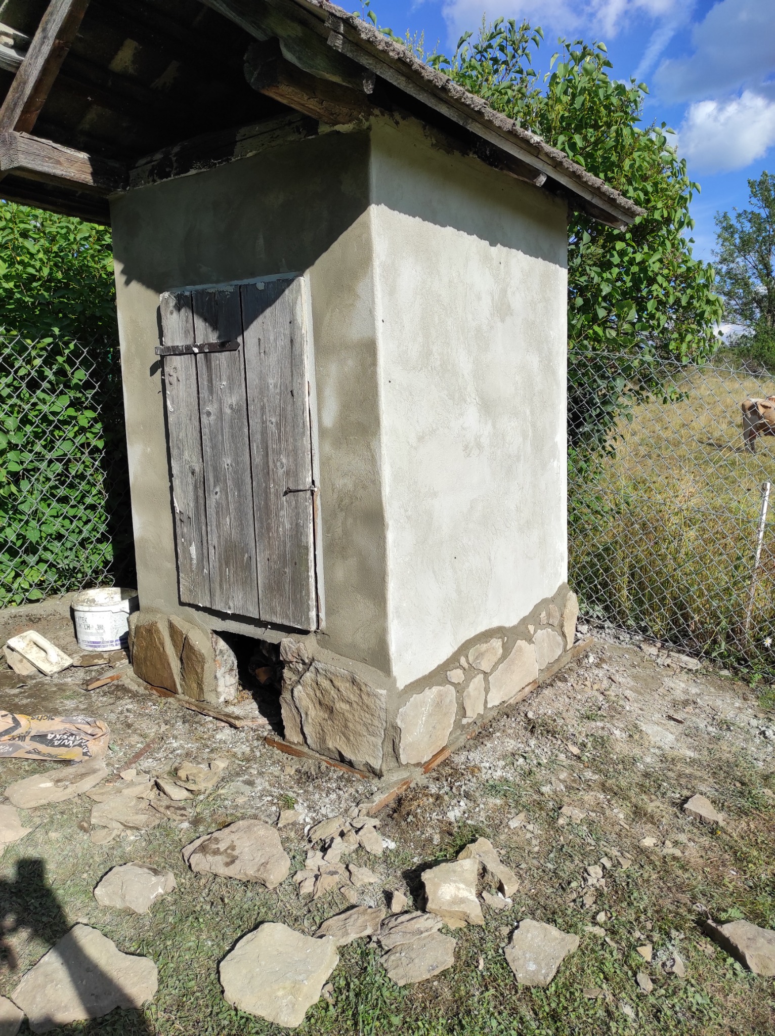 Mała, otynkowana budowla z drewnianymi drzwiami, otoczona kamienną podstawą i siatką ogrodzeniową, z widocznymi śladami prac budowlanych i workiem zaprawy.