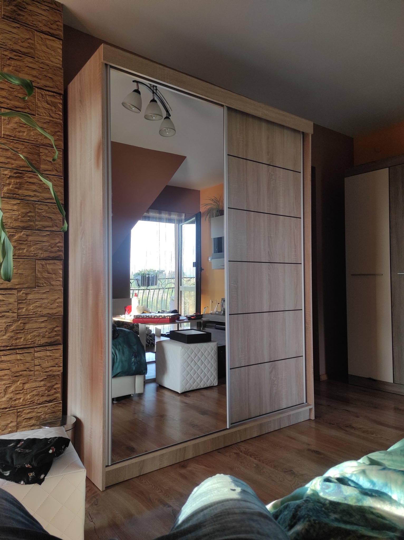 Szafa przesuwna z lustrem odbijająca wnętrze pokoju z wyjściem na balkon, widoczna część podłogi z paneli i dekoracyjna ściana z cegieł.