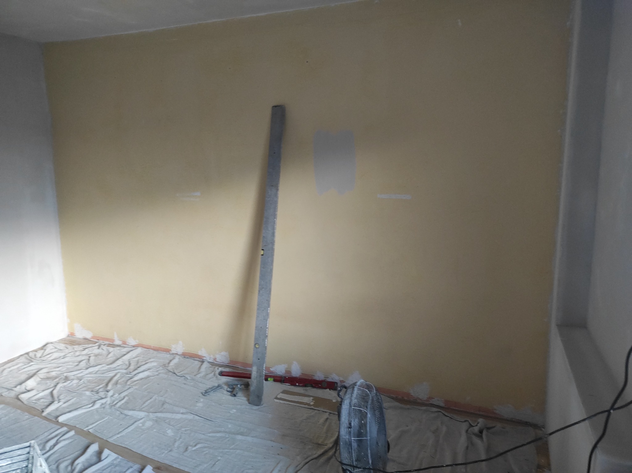 Pokój w trakcie przygotowania do malowania, z żółtą ścianą, częściowo zagruntowaną, zabezpieczoną podłogą i narzędziami malarskimi opartymi o ścianę.