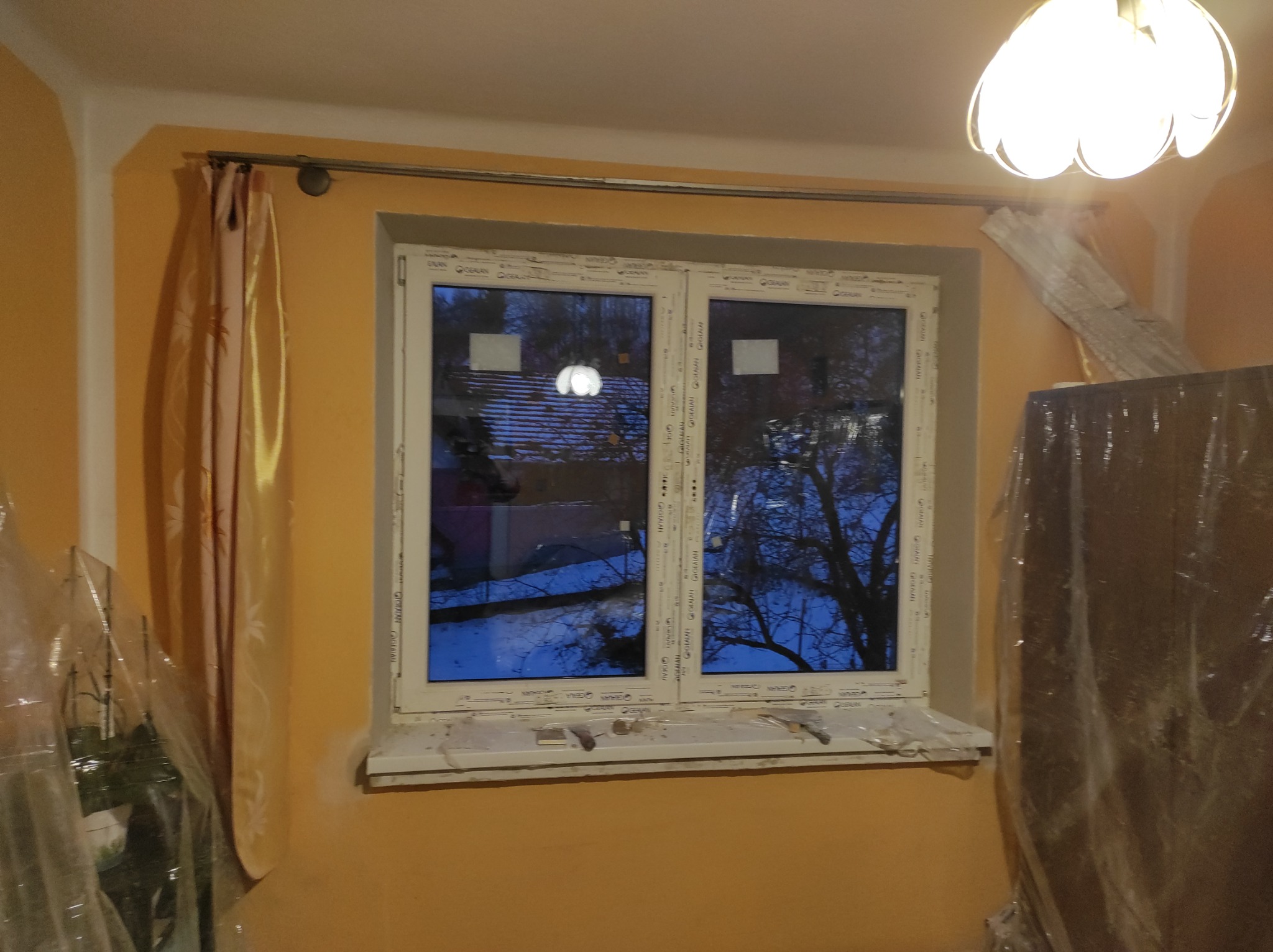 Pokój w trakcie remontu: pomarańczowe ściany, okno PCV z widokiem na drzewa, zasłonięte meble folią ochronną, widoczny karnisz i lampa sufitowa.