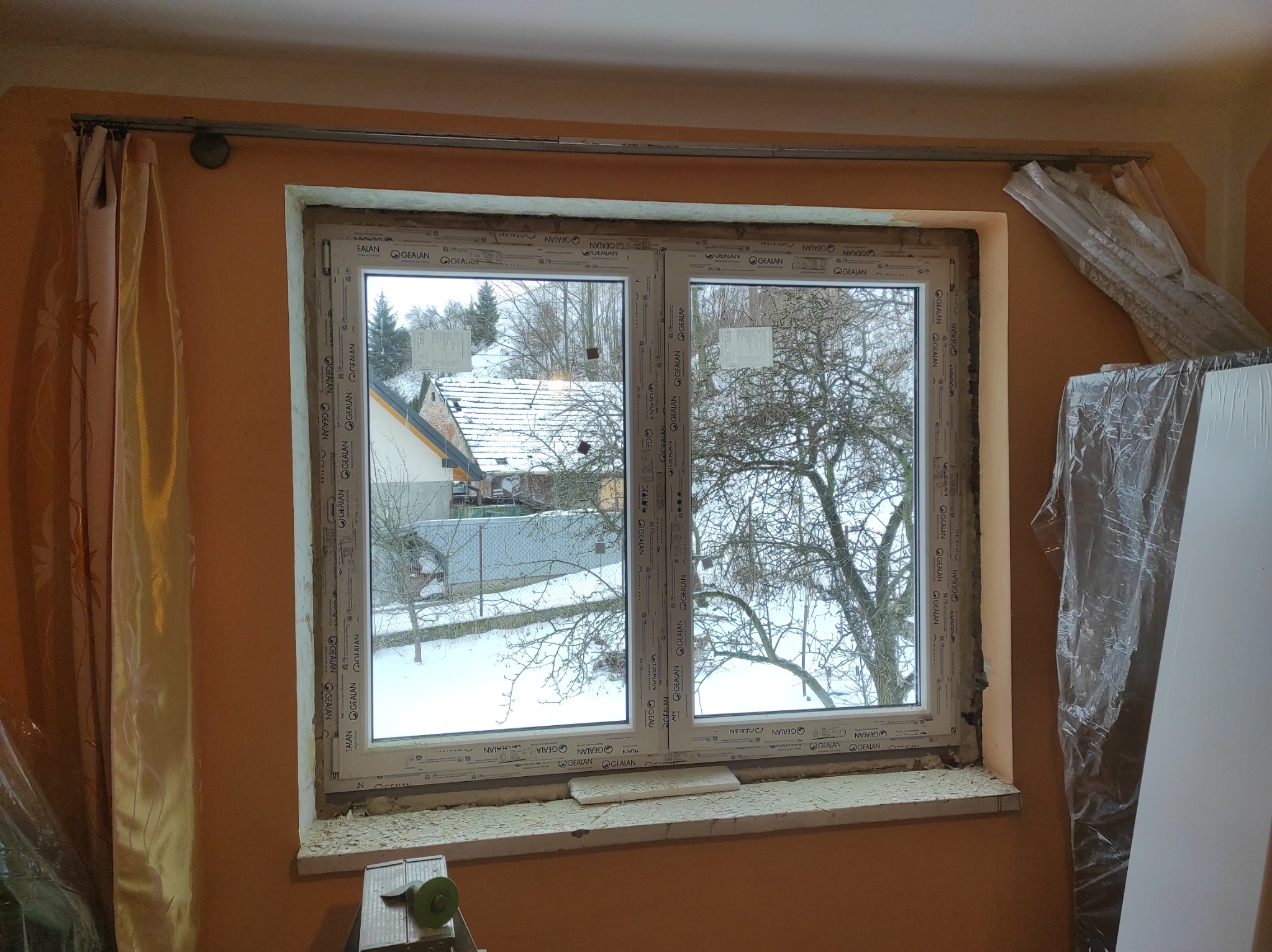 Montaż okna PCV z widokiem na zimowy krajobraz, ślady pianki montażowej na parapecie, częściowo zasłonięte zasłoną i folią ochronną.