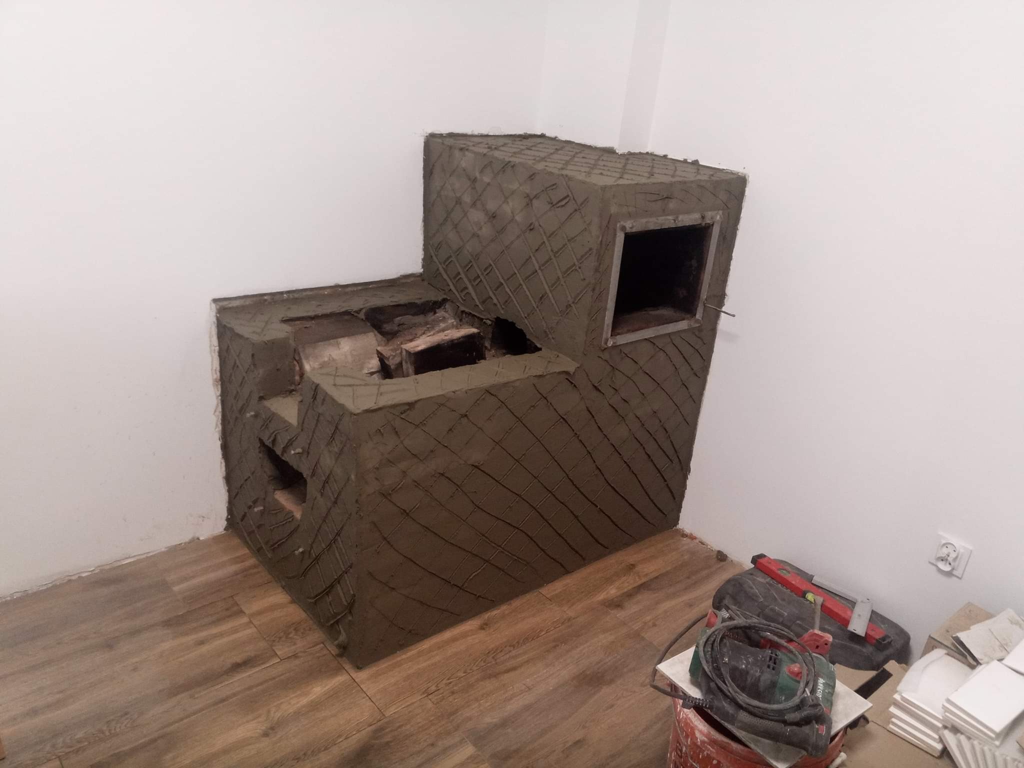 Surowy kominek obłożony siatką i świeżo nałożoną warstwą szarej zaprawy, widok z boku na podłodze z paneli, w tle biała ściana i gniazdko elektryczne, obok narzędzia i wiadro.