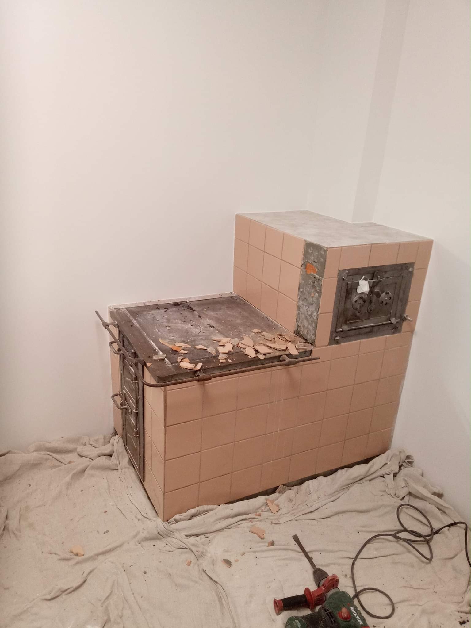 Demontaż kafli pieca kaflowego, widoczne fragmenty odspojonych płytek, zabezpieczona podłoga z narzędziami do demontażu w tle, ściany przygotowane do dalszych prac.