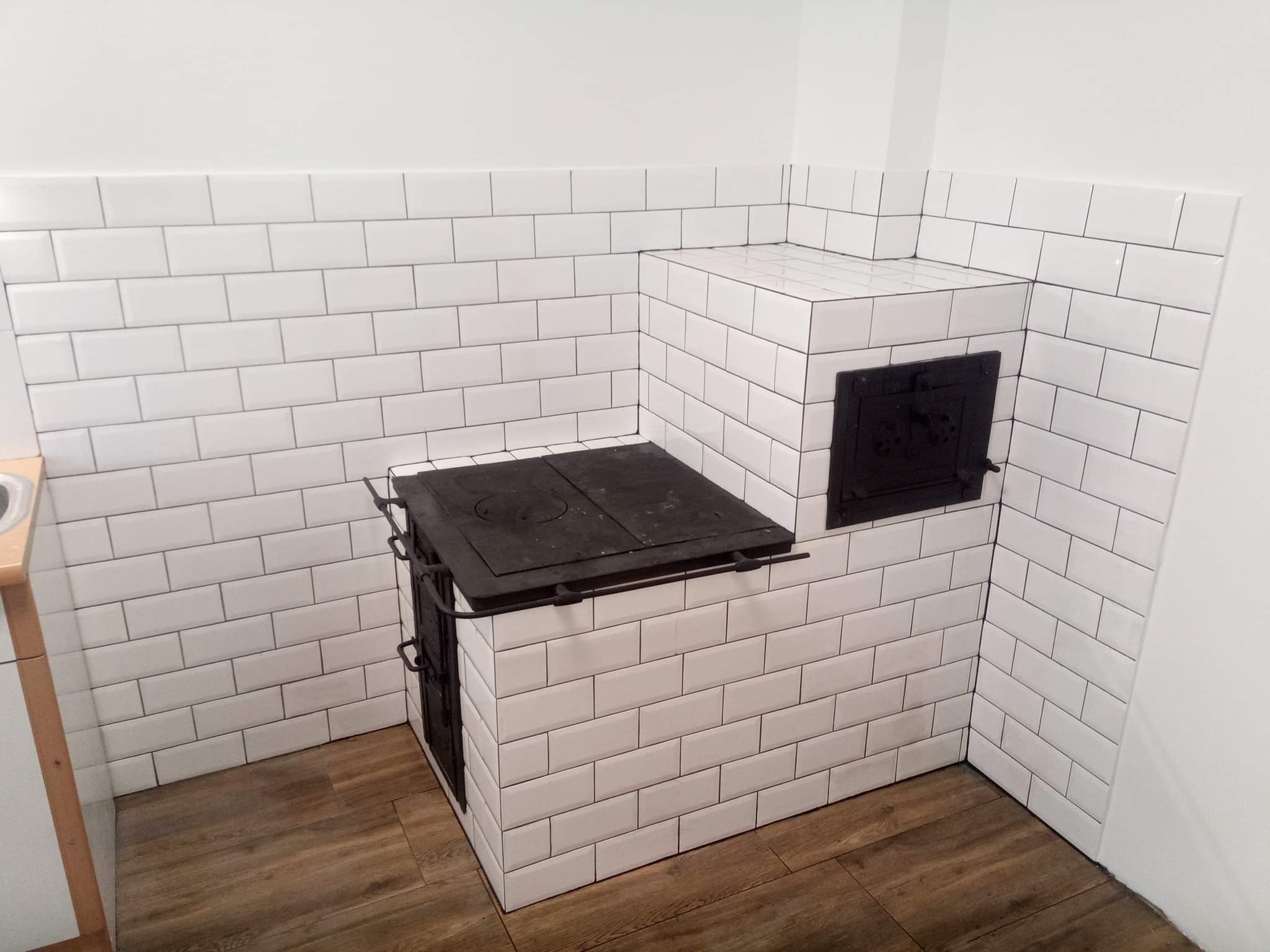 Kuchenny piec kaflowy z czarnymi żeliwnymi drzwiczkami i płytą grzewczą, obłożony białymi, prostokątnymi płytkami ceramicznymi z ciemną fugą, na podłodze panele imitujące drewno.