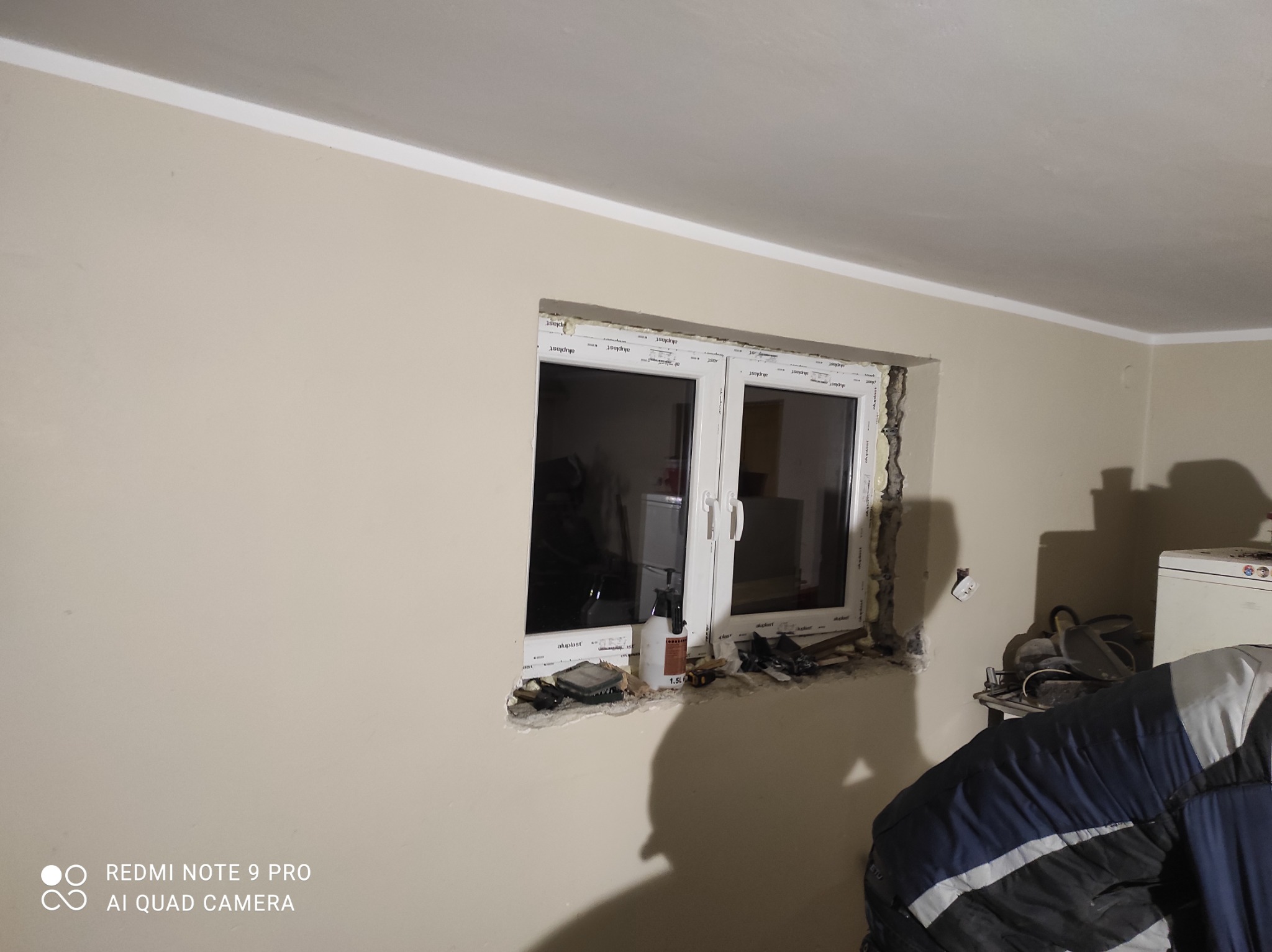 Montaż okna PCV w ścianie, widoczne narzędzia i piana montażowa, etap prac wykończeniowych w jasnym pomieszczeniu.