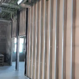 Montaż ścian działowych z płyty karton-gips na metalowym stelażu, widoczne instalacje wentylacyjne pod sufitem oraz rolka materiału izolacyjnego przy ścianie.