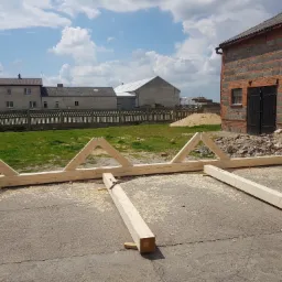 Konstrukcja drewniana więźby dachowej, wstępny montaż na betonowym podłożu, widoczne elementy trójkątne i poziome belki, w tle budynki gospodarcze i błękitne niebo z chmurami.