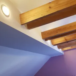 Skośny sufit poddasza z widocznymi drewnianymi belkami, oświetleniem punktowym i pomalowanymi ścianami w odcieniach fioletu i szarości.